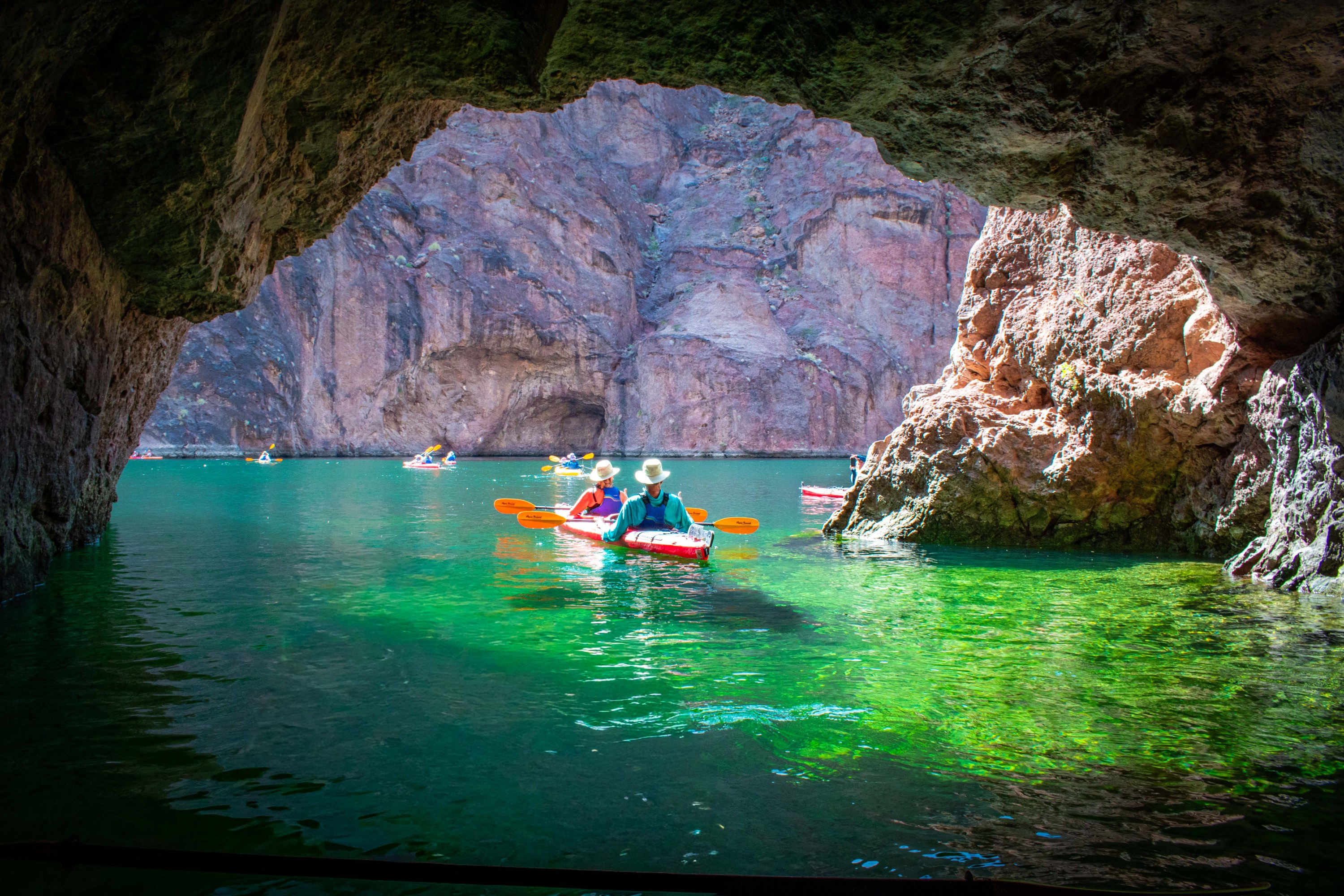Kayakers paddle through cave with rocky walls, green water, and daylight in the background.